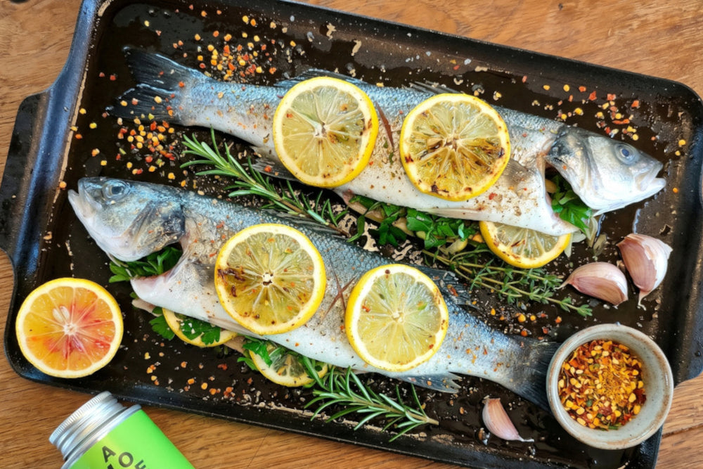 Zwei ganze Fische liegen mit Zitronenscheiben, Rosmarin, Knoblauch und Gewürzen auf einem schwarzen Grillblech