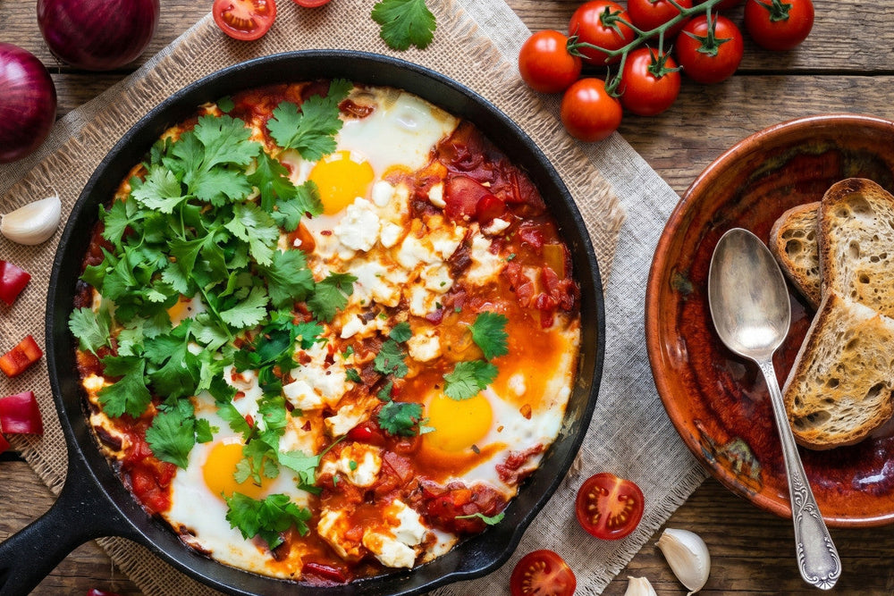 Eine Shakshuka mit Eiern, Tomatensauce, Feta und Koriander steht in einer Pfanne neben Tomaten, Brot und Gewürzen