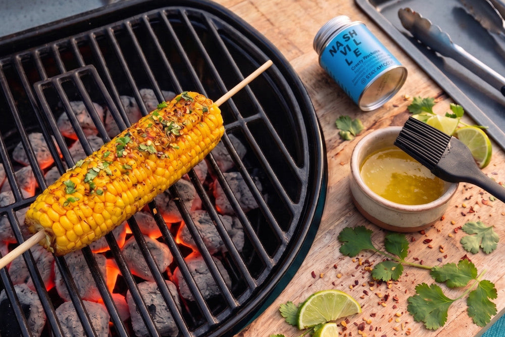 Ein Maiskolben mit würziger Crusty Nashville Butter liegt auf dem Grill; daneben sind Butter, Pinsel und Gewürzdose zu sehen