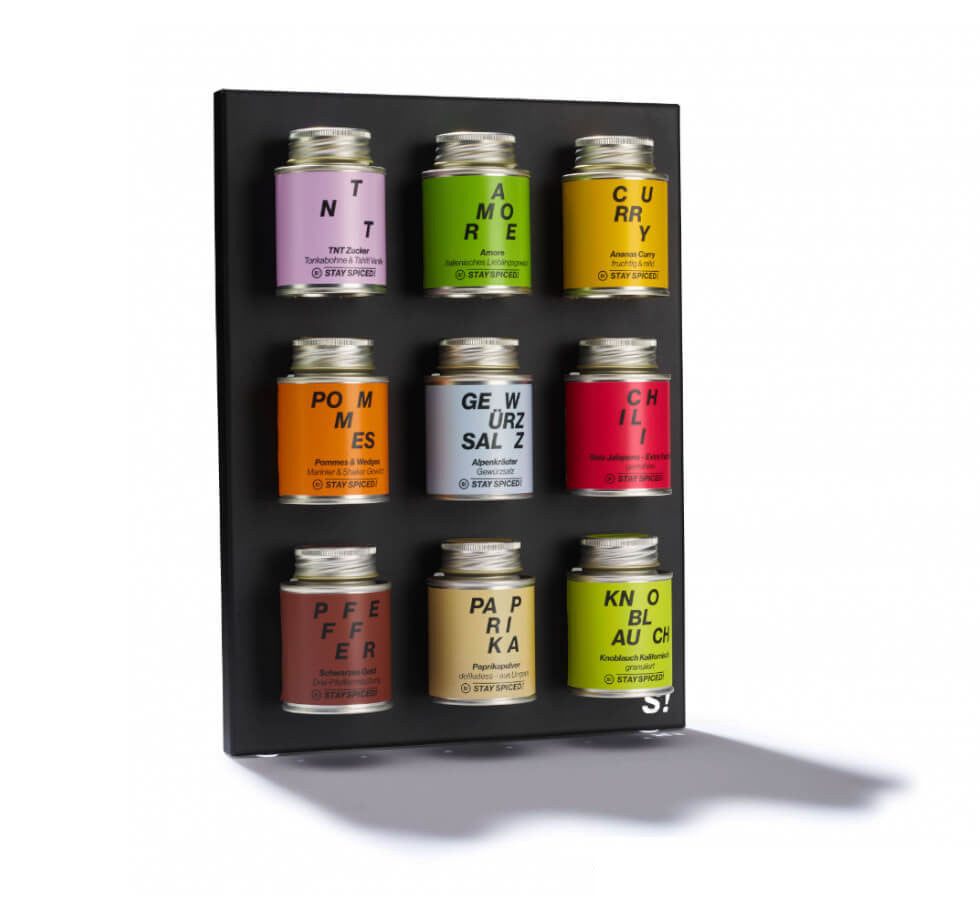An der Wand montierter schwarzer Gewürzständer bestückt mit neun bunten STAY SPICED ! Gewürzdosen