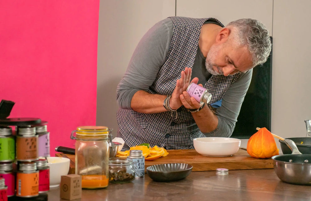 Ein passionierter Koch verfeinert ein Gericht mit einer STAY SPICED! Gewürzdose in einer modernen Küche. Im Hintergrund ist ein stilvolles Gewürzregal für die optimale Aufbewahrung von Chefkoch-Gewürze zu sehen.