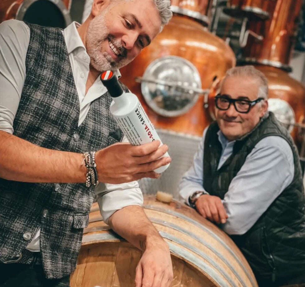 Roland Trettl und Hans Reisetbauer präsentieren stolz ihre handgefertigte Bio-Soja Sauce vor Holzfässern in der traditionellen Qualitätsbrennerei